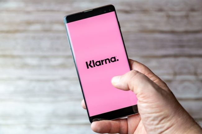 Hand som håller i telefon med rosa bakgrund och texten Klarna i svart.